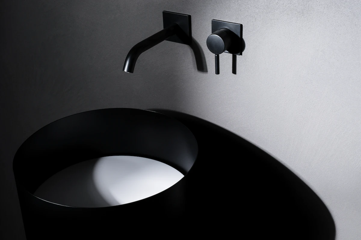 lavabo cilindrico freestanding in acciaio nero con rubinetto e manopola a muro, inserito in un contesto di design minimal
