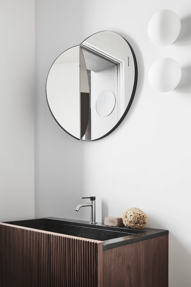 bagno moderno con lavandino in pietra scura e legno a doghe, specchio rotondo dalla forma asimmetrica ed accessori da bagno