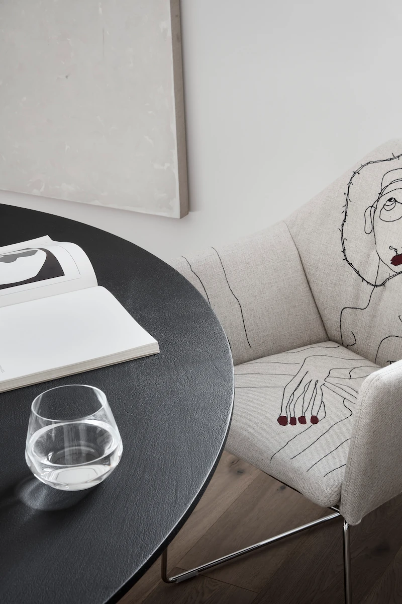 dettaglio di sedia imbottita con ricami artistici accanto a un tavolo rotondo nero, con bicchiere d’acqua e libro aperto