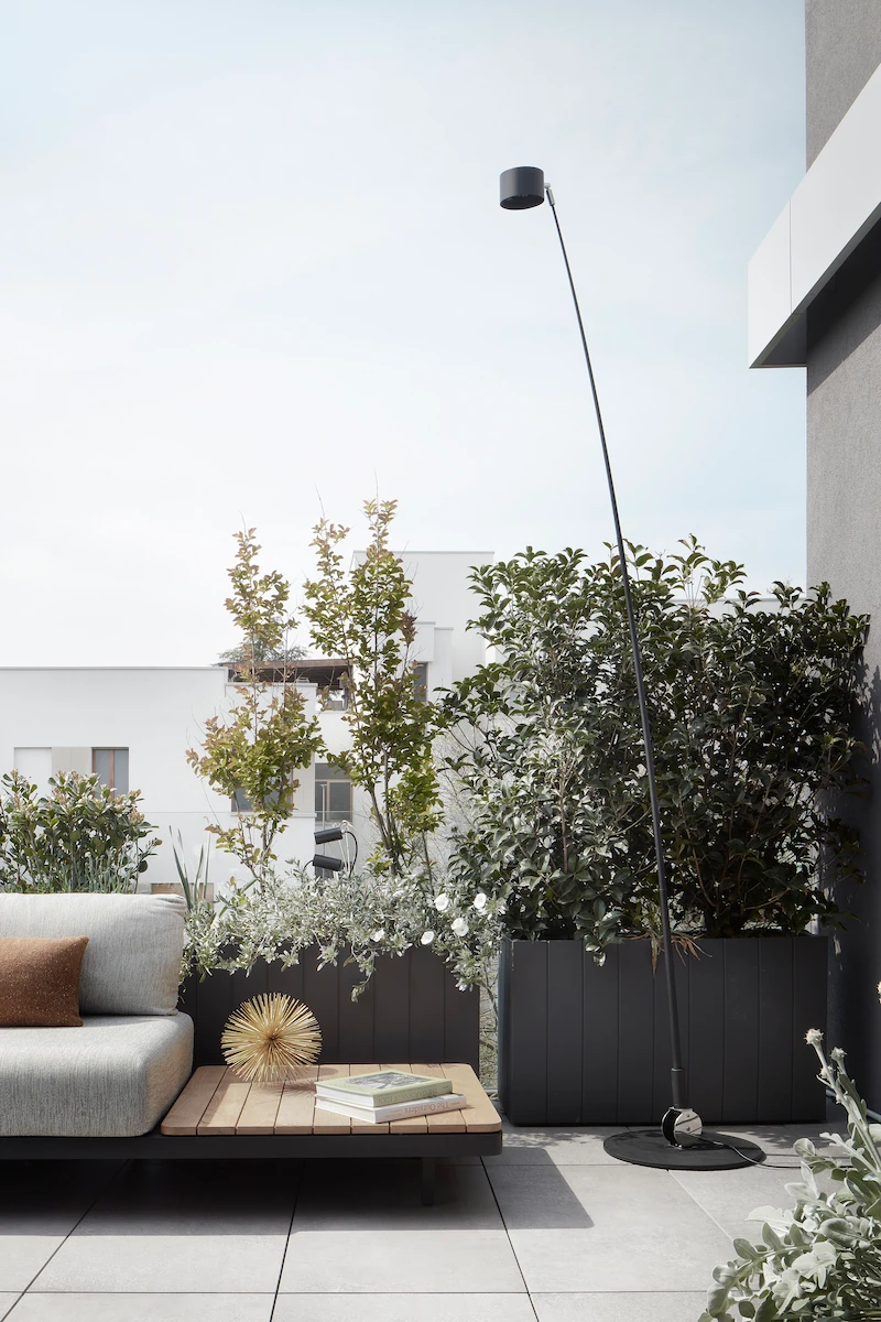 angolo di una terrazza moderna con divano grigio, tavolino in legno e una lampada da esterno ad arco, circondato da piante