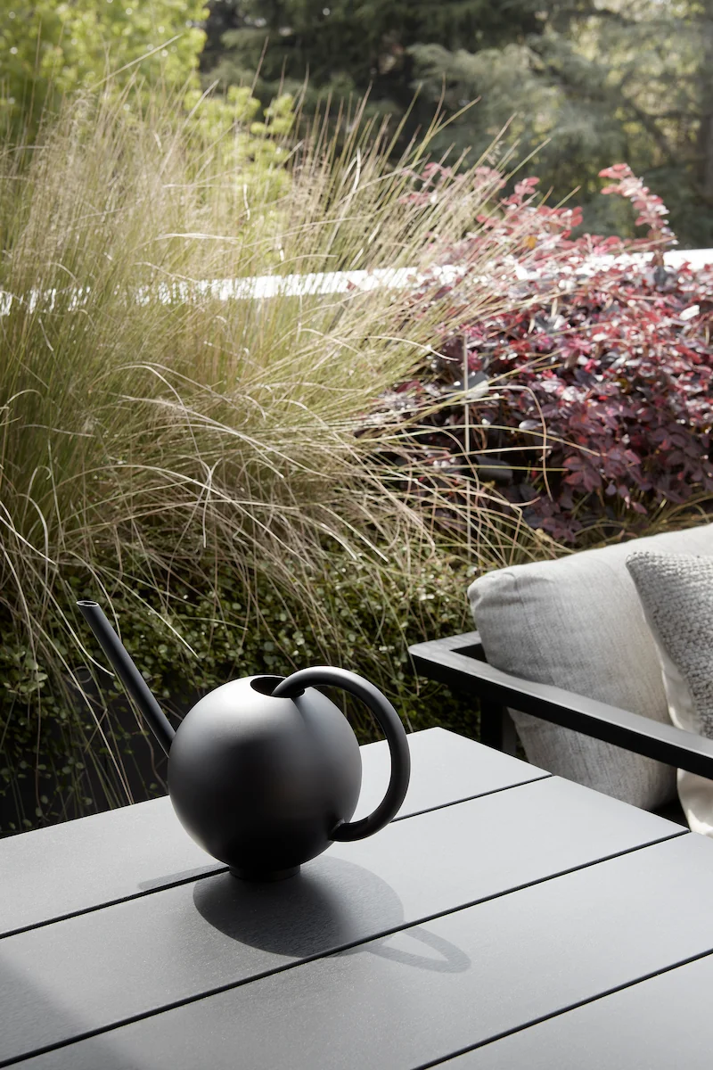 innaffiatoio nero dal design minimalista su un tavolo da esterno, con piante rigogliose sullo sfondo